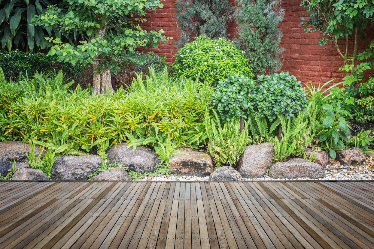 Jardin paysager extérieur avec terrasse en bois composite, bordure de pierres naturelles et plantation mixte de fougères, arbustes persistants et conifères nains contre mur de briques rouges